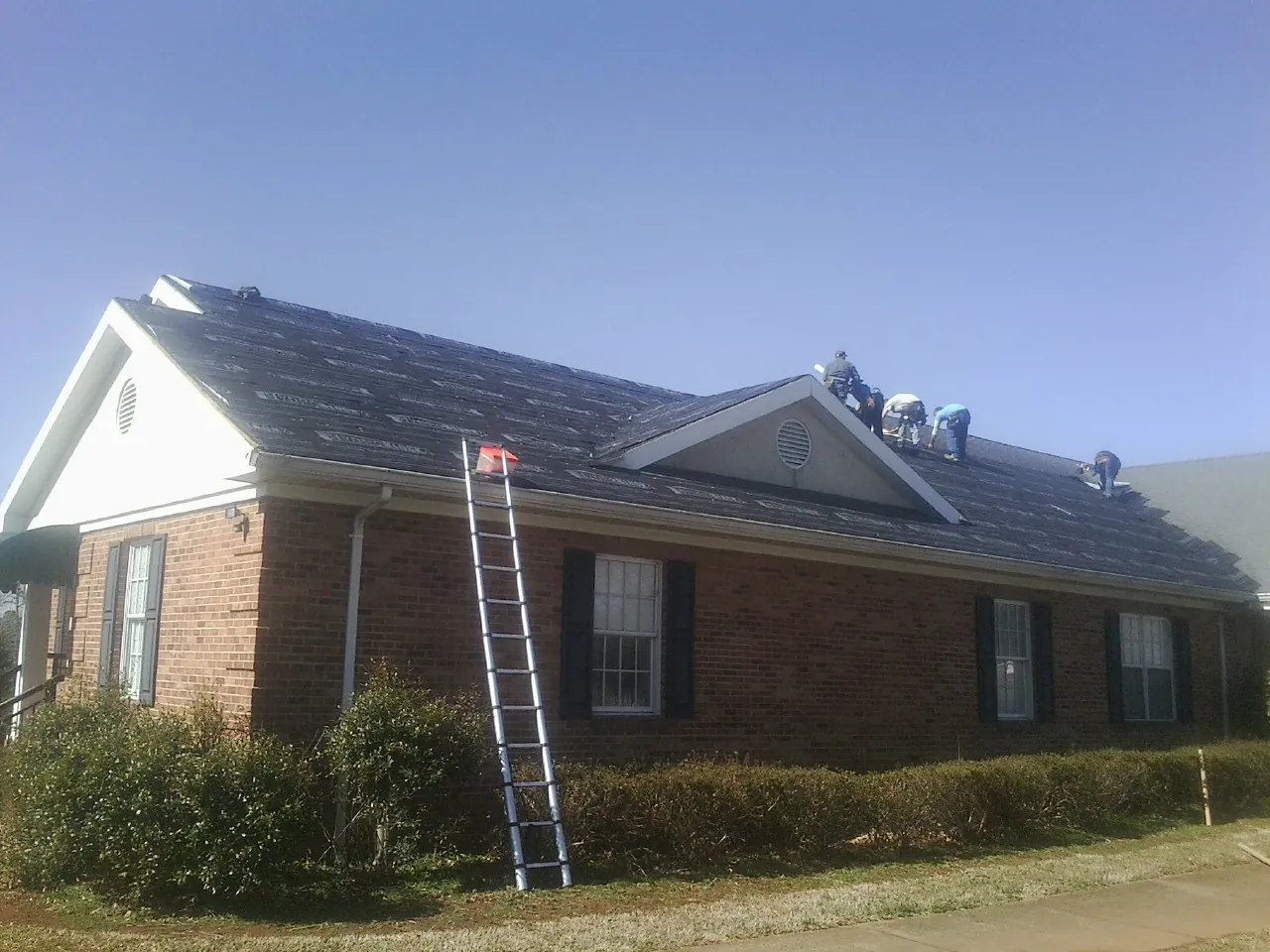 Crew tearing shingles off a brick ranch roof with a ladder set — Emergency Roof Repair work by our crew in Arcanum