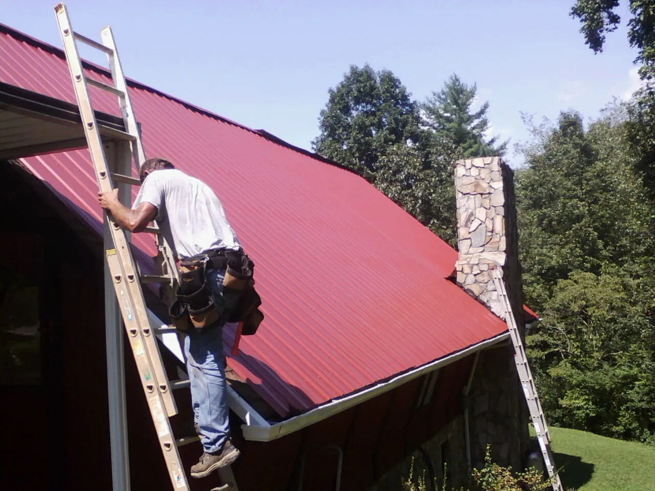 Roofer on a ladder beside a red metal roof and stone chimney — Soffit & Fascia Repair by Roofing Brothers in Arcanum
