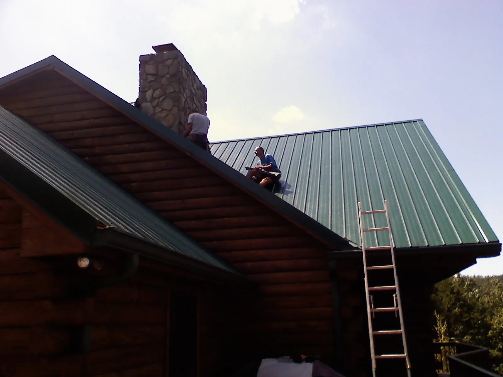 Roofers installing green metal panels beside a stone chimney on a log home in Arcanum